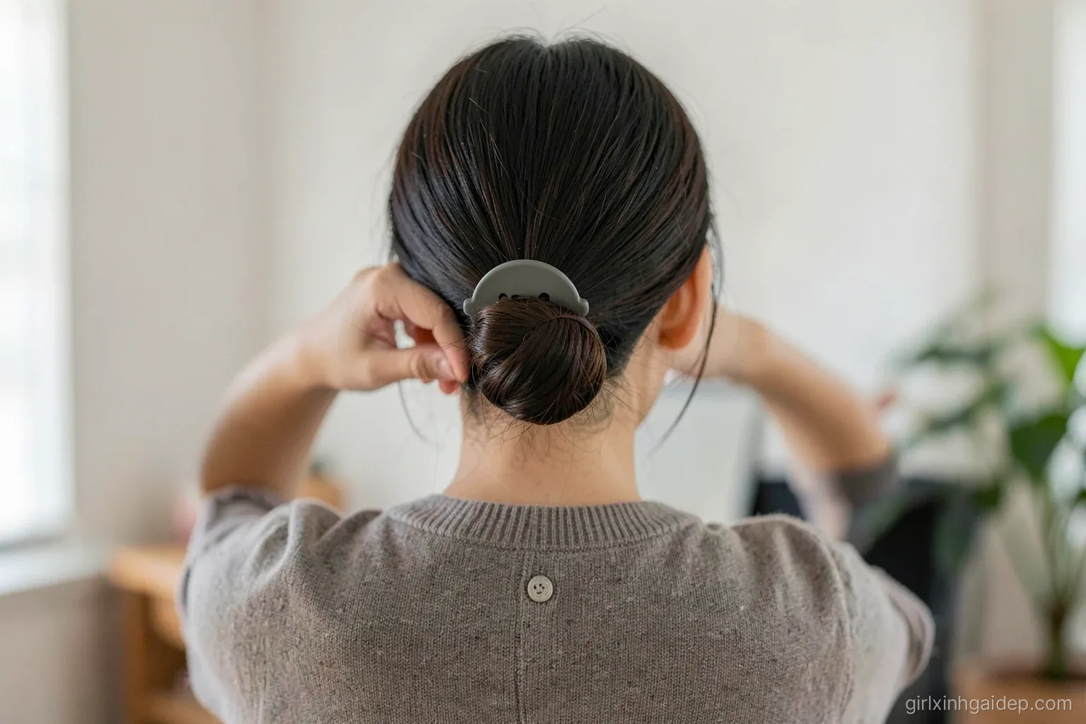 Close-up from behind of an adult Vietnamese woman twisting her hair into a low bun secured with a large matte claw clip, office cardigan, soft indoor daylight