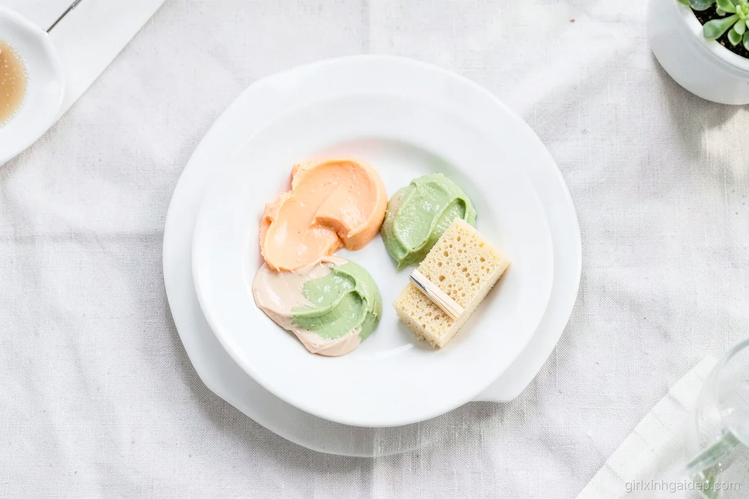 Top-down view of a white ceramic plate holding dabs of peach, green and beige creams beside a tiny flat brush and a damp sponge, plain linen tablecloth, bright diffused light